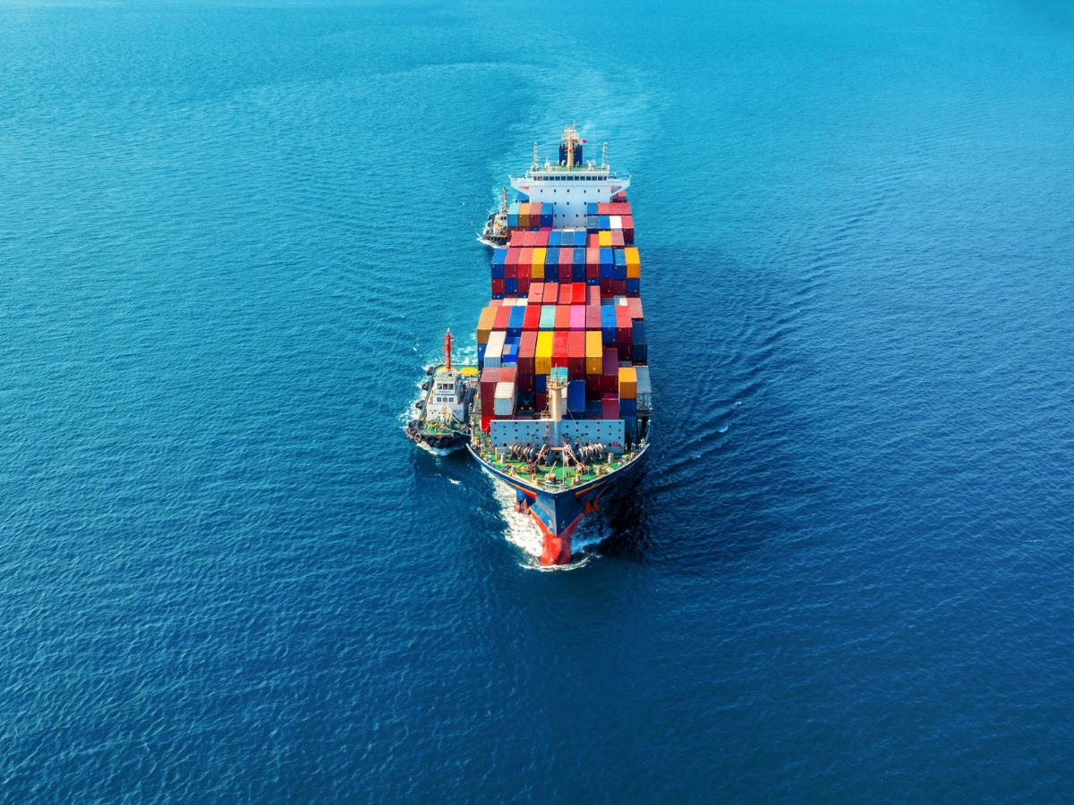 Aerial view of a container ship loaded with colorful shipping containers, escorted by two tugboats across calm deep-blue open water.