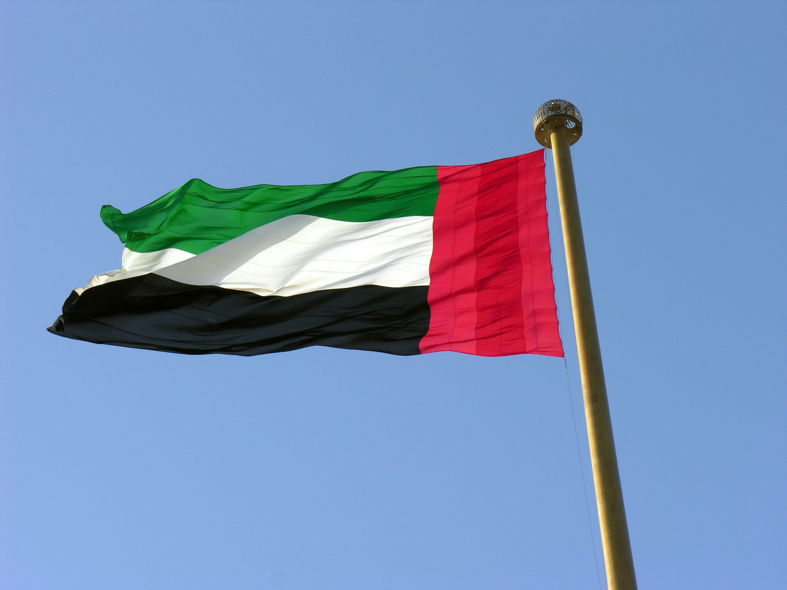 The national flag of the United Arab Emirates fluttering in the wind on a golden flagpole against a clear blue sky.