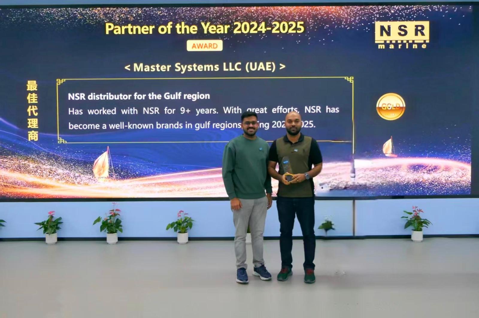Two men standing in front of a large screen displaying "Partner of the Year 2024-2025 Award" for Master Systems LLC (UAE), one holding the award trophy